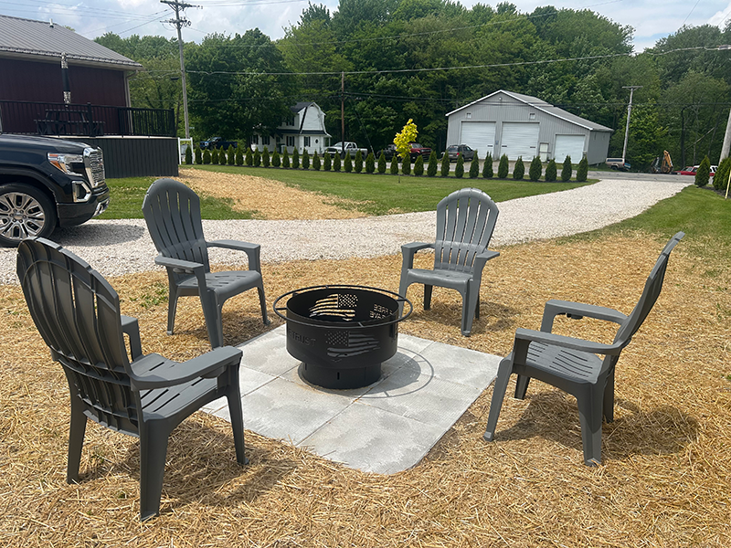 firepit A fire pit on a square concrete slab surrounded by four grey deck chairs in an outdoor area with trees in the background.